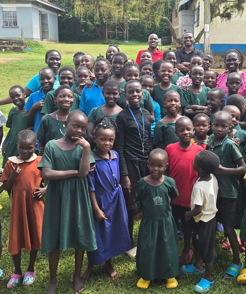 Smiling schoolchildren, many in green uniforms, gathered on a lawn in front of their school.