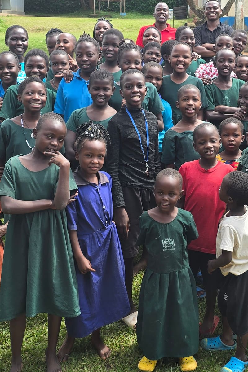 Smiling schoolchildren, many in green uniforms, stand close together on a lawn.