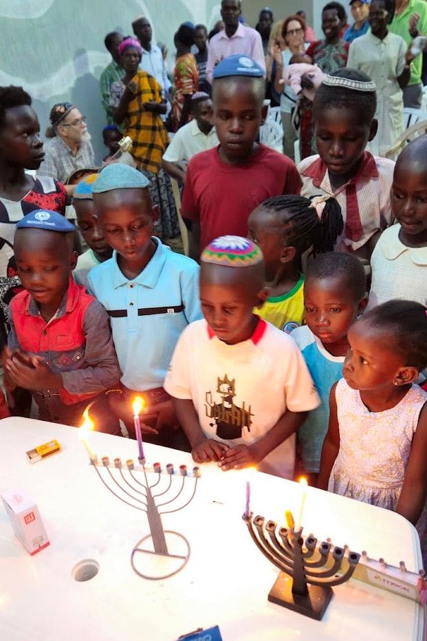 Children, some wearing kippot, gather at a table at night to watch Hanukkah candles burn on two menorahs.