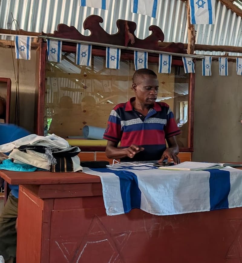 A man stands at a table beside a stack of handmade textiles, under a string of Israeli-flag bunting.
